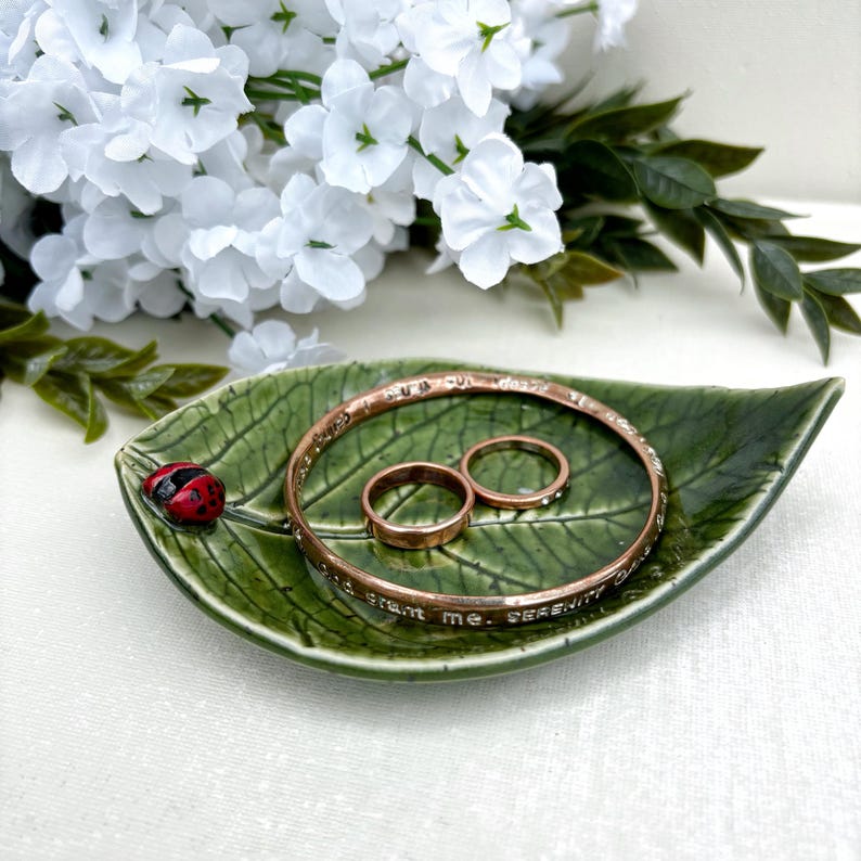 Pu&ograve; includere: Un piattino in ceramica verde a forma di foglia con una piccola statuetta di coccinella rossa e nera sul bordo. All'interno del piattino poggia un sottile bracciale in metallo color rame inciso con testo e due anelli semplici abbinati.