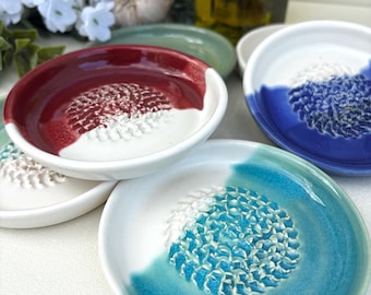 May include: A set of four round ceramic grater plates, each featuring a central textured area for grating surrounded by a smooth, coloured rim. The plates are glazed in red, blue, turquoise, and white, with a white base and raised grating teeth.