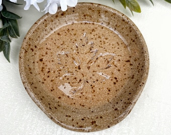 May include: A round, tan ceramic spoon rest with a speckled finish and a glossy glaze. The centre of the dish features an embossed maple leaf design. The ceramic surface has a light brown base colour with darker brown speckles throughout the piece.