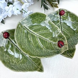 May include: Three ceramic dishes shaped like green leaves with detailed veins and a glossy finish. Each leaf features a small, three-dimensional red ladybug with black spots attached to the surface, creating a textured and realistic appearance.