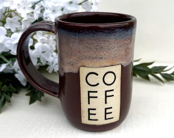 May include: A ceramic mug with a dark maroon glaze and a textured, light brown band near the rim. A rectangular cream-coloured panel on the front features the word COFFEE stacked vertically in black sans-serif capital letters. The mug has a handle.