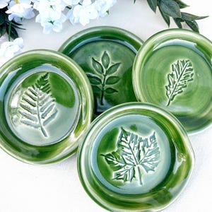 May include: A set of four round ceramic dishes with a glossy green glaze. Each dish features a unique, embossed botanical impression in the center, including a fern, a compound leaf, an oak leaf, and a maple leaf, all with raised edges.