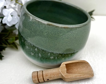 May include: A round ceramic bowl with a smooth, glossy finish featuring a gradient of sage green at the top transitioning into a darker forest green and white at the base. A small, light-coloured wooden scoop with a grooved handle rests in front.
