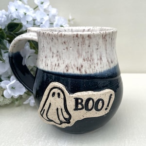 May include: A ceramic mug with a white and speckled top, a dark blue base, and a handle. A white ghost and the word "BOO!" are on the front. The mug is likely for Halloween.