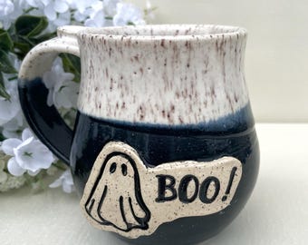 May include: A ceramic mug with a white and speckled top, a dark blue base, and a handle. A white ghost and the word "BOO!" are on the front. The mug is likely for Halloween.