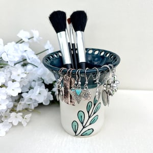 May include: A ceramic makeup brush holder with a dark teal rim and a white base, adorned with a green leaf design. The holder contains makeup brushes and a variety of earrings. The earrings are silver, gold, and turquoise.