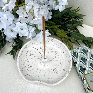May include: A round, white ceramic incense holder with a speckled black pattern and a central upright post. A thin brown incense stick is inserted into the holder. Beside it sits a box with a green and white geometric fan-shaped pattern.