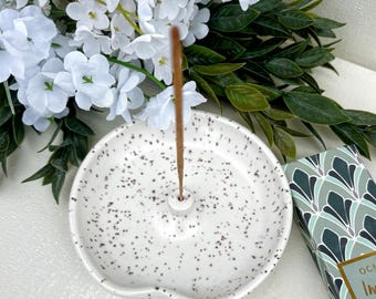 May include: A round, white ceramic incense holder with a speckled black pattern and a central upright post. A thin brown incense stick is inserted into the holder. Beside it sits a box with a green and white geometric fan-shaped pattern.
