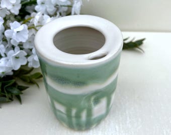 May include: A ceramic cup with a smooth white rim and a cylindrical body featuring a sage green and white drip glaze pattern. The vessel has a wide circular opening at the top and a small, integrated horizontal slit near the rim edge.
