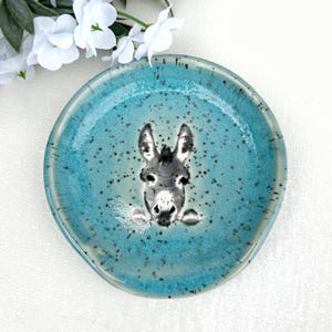 Peut inclure: Une coupelle en céramique ronde avec un émail turquoise moucheté et une illustration centrale en noir et blanc représentant une tête d'âne. L'âne est représenté de face avec les oreilles dressées, centré sur la surface brillante et texturée.