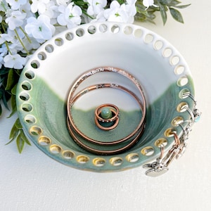 May include: A ceramic bowl with a white interior and a sage green exterior featuring a perforated rim. The bowl contains two copper-toned metal bangles and a small ring resting on a central post, with silver-toned charms hanging from the edge.