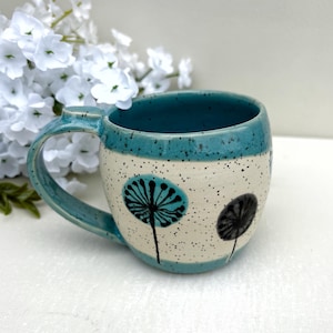 Può includere: Una tazza in ceramica con corpo color crema maculato e bordo e manico color verde acqua. L'esterno presenta motivi a dente di leone neri dipinti a mano. La tazza ha una forma arrotondata e una finitura liscia e lucida, che mostra un netto contrasto tra gli accenti verde acqua e la base chiara maculata.