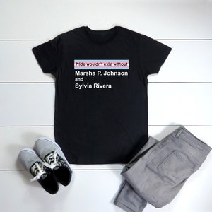 Puede incluir: Camiseta negra con el texto "Pride wouldn't exist without Marsha P. Johnson and Sylvia Rivera" impreso en ella. El texto está enmarcado por una bandera del orgullo transgénero rosa, azul y blanca.
