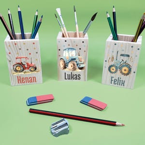 Könnte beinhalten: Drei Holz-Stifthalter mit Traktorillustrationen und den Namen "Henan", "Lukas" und "Felix". Jeder Halter enthält Bleistifte und Pinsel. Außerdem liegen zwei rosa-blaue Radiergummis, ein Bleistift und ein Spitzer auf einer grünen Oberfläche.