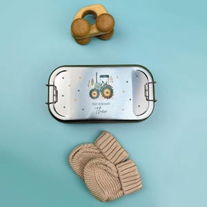May include: A silver lunchbox with a blue tractor design and the text "Hier kr&uuml;melt Theo". The lunchbox is next to a pair of beige knitted mittens and a wooden toy.