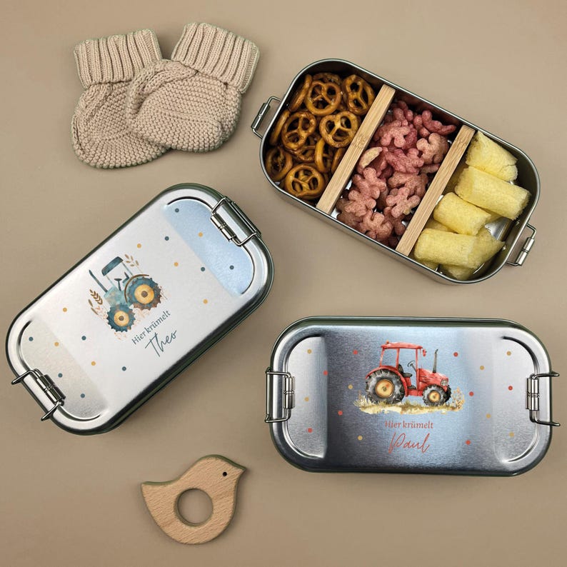 May include: Two stainless steel lunchboxes with a tractor design and the text "Hier brummelt Theo" and "Hier brummelt Paul". The lunchboxes are filled with snacks like pretzels, puffed rice cereal, and cheese puffs.