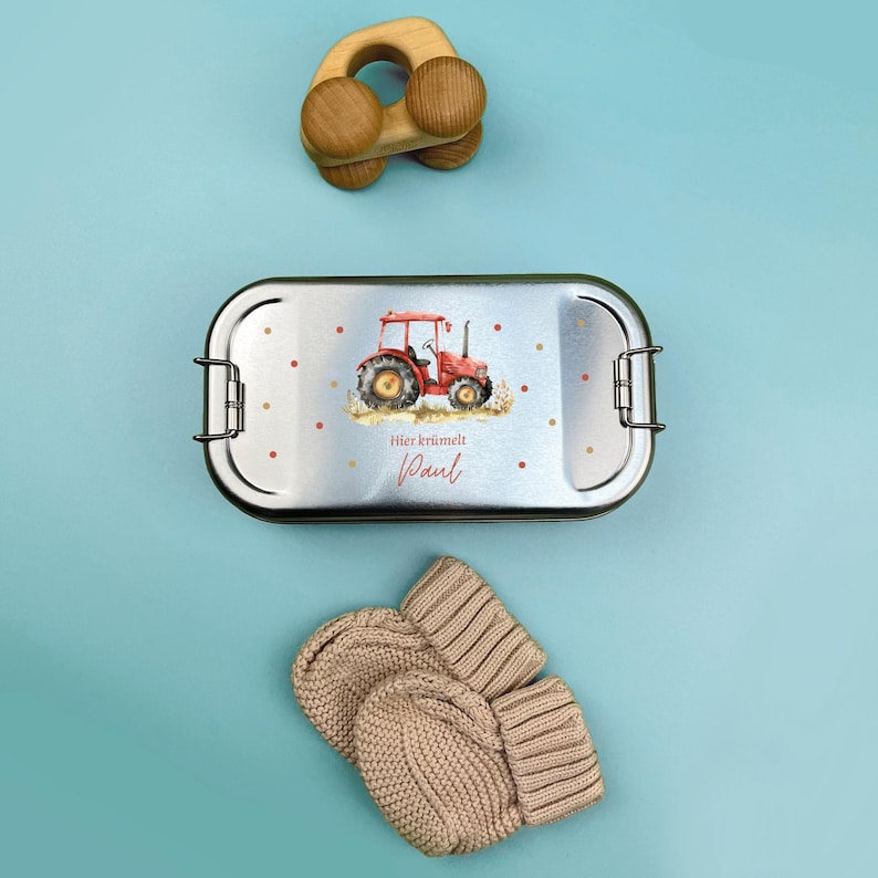 May include: A silver lunchbox with a red tractor and the text "Hier kr&uuml;melt Paul" on the lid. The lunchbox is next to a pair of beige knitted mittens.