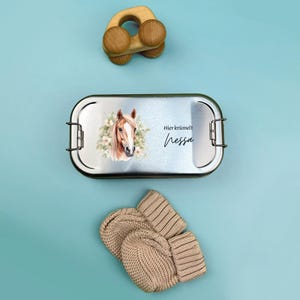 K&ouml;nnte beinhalten: Eine silberne Lunchbox mit einer Pferdeillustration und dem Text "Hier kr&uuml;melt Nessa". Die Lunchbox liegt neben einem Paar beigefarbener Strickhandschuhe und einem Holzspielzeug.