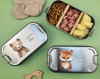 May include: Two stainless steel lunch boxes with animal illustrations and the text "Hier krümelt Ben" and "Hier krümelt Fred". The lunch boxes are filled with snacks like pretzels, puffed rice, and cheese puffs.
