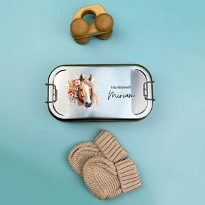 K&ouml;nnte beinhalten: Eine silberne Brotdose mit einer Pferdeillustration und dem Text "Hier kr&uuml;melt Miriam". Die Brotdose liegt neben einem Paar beigefarbener Strickhandschuhe und einem Holzspielzeug.