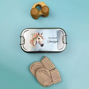 K&ouml;nnte beinhalten: Eine silberne Lunchbox mit einem wei&szlig;en Pferd und einem Blumenmuster. Die Lunchbox hat den Text "Hier kr&uuml;melt Vanessa" drauf.