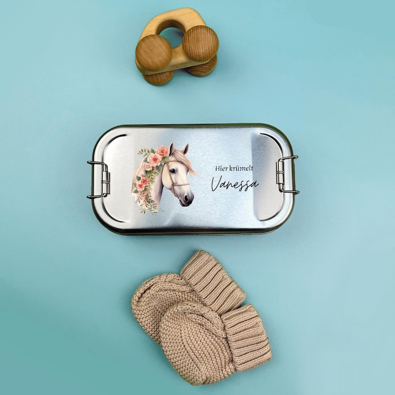 K&ouml;nnte beinhalten: Eine silberne Lunchbox mit einem wei&szlig;en Pferd und einem Blumenmuster. Die Lunchbox hat den Text "Hier kr&uuml;melt Vanessa" drauf.