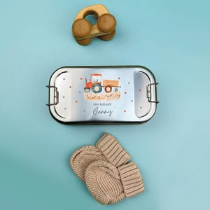 May include: A silver lunchbox with a red tractor and the text "Hier kr&uuml;melt Benny" on the front. The lunchbox is next to a wooden toy car and a pair of beige knitted mittens.