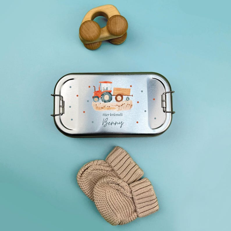 May include: A silver lunchbox with a red tractor and the text "Hier kr&uuml;melt Benny" on the front. The lunchbox is next to a wooden toy car and a pair of beige knitted mittens.