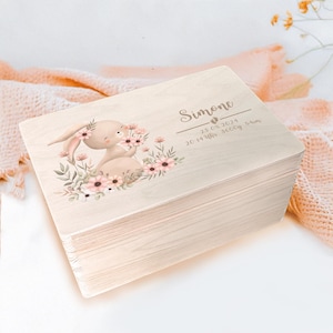Könnte beinhalten: Eine Holz-Erinnerungskiste mit hellbrauner Oberfläche. Die Kiste zeigt eine niedliche Illustration eines Hasen, der Blumen hält, und den Text "Simone 23.08.2024 20:14 Uhr 3600g 54cm".