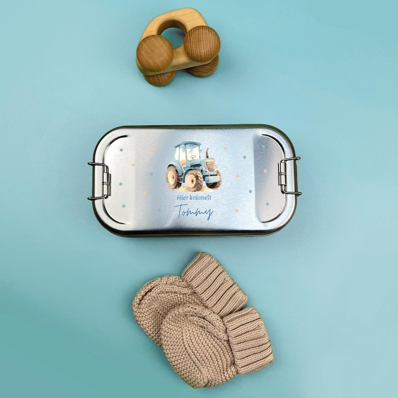 May include: A silver lunchbox with a blue tractor design and the text "Hier kr&uuml;melt Tommy". The lunchbox is on a light blue surface with a wooden toy car.