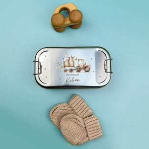 May include: A silver lunchbox with a colorful illustration of a tractor and animals. The lunchbox has the text "Hier kr&uuml;melt Kilian" printed on it.