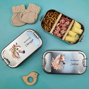 K&ouml;nnte beinhalten: Zwei Edelstahl-Lunchboxen mit einem Pferde- und Blumendesign. Die Lunchboxen haben den Text "Hier krimelt Vanessa" und "Hier krimelt Leonie" aufgedruckt. Die Lunchboxen befinden sich auf einer blauen Oberfl&auml;che mit einem h&ouml;lzernen Spielzeugvogel.
