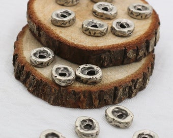 May include: A collection of silver-colored, textured metal beads with a central hole, scattered on two rustic wooden slices. The beads have an irregular, organic shape, suggesting a handmade or vintage aesthetic. The background is a plain white surface.