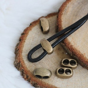 May include: A close-up of a black leather cord with antique gold metal cord ends and decorative metal beads. The cord and beads are on a wooden surface.