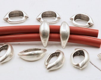Könnte beinhalten: Silberfarbene Metall-Schmuckteile, darunter ovale Glieder und längliche, tropfenförmige Perlen. Die Teile sind auf einer weißen Oberfläche mit zwei roten, zylindrischen Stäben angeordnet. Das Metall hat eine matte Oberfläche.