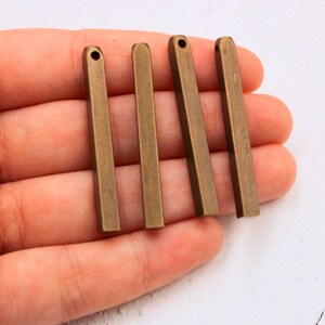 May include: Five antique gold-toned metal bar pendants with a smooth, slightly textured finish. Each pendant has a hole at the top for attaching a chain or string.