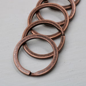 May include: Close-up of several antique copper-colored key rings. The rings are circular with a split for attaching keys. The metal has a textured, aged appearance, suggesting a vintage or rustic aesthetic. The rings are arranged in a slightly overlapping pattern.
