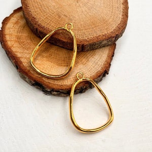 Puede incluir: Dos colgantes de metal de color dorado en forma de &oacute;valo irregular. Los colgantes est&aacute;n sobre una superficie de madera.