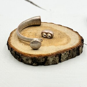 Könnte beinhalten: Ein silbernes Armreif-Armband mit abgerundetem Ende und einem separaten silbernen Verbindungsteil mit zwei Löchern. Das Armband und der Verbinder werden auf einer Holzscheibe präsentiert.