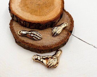 May include: Three gold-toned metal charms in the shape of hands. The charms are on a wooden surface.