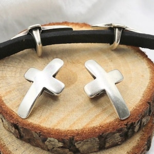 May include: Two silver cross charms with a smooth, slightly textured surface. The charms are displayed on a wooden surface.