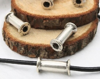 May include: Silver metal beads in the shape of spools, with a hole in the center of each bead. The beads are arranged on a wooden surface and one bead is strung on a black leather cord.