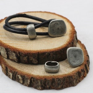 May include: A black leather bracelet with silver metal accents. The bracelet has two silver metal beads and a silver metal clasp. The bracelet is displayed on a wooden surface.