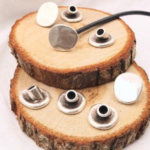 May include: A set of silver metal eyelets for leather crafting. The eyelets are round with a hole in the center. They are arranged on a wooden surface.