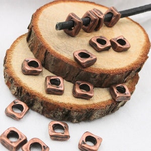 May include: A group of copper-coloured square beads with a hole in the centre. The beads are arranged on a wooden surface and some are strung on a black cord.