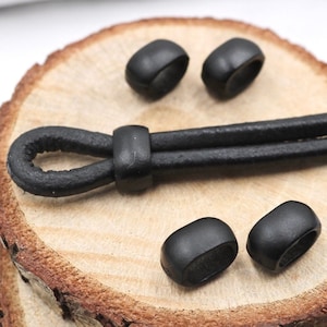 May include: Five black cord end stoppers, four are round and one is oval, on a wooden surface. The stoppers are used to finish the ends of cords and straps.