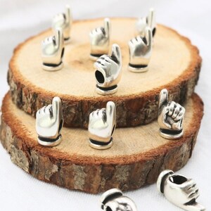 May include: Silver charm beads in the shape of a pointing finger. The beads are arranged on a wooden surface.