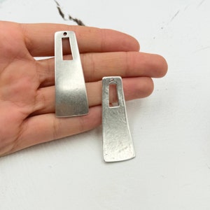 May include: Two silver-toned metal pendant charms with a rectangular shape and a cut-out rectangle in the center. The charms have a textured surface.