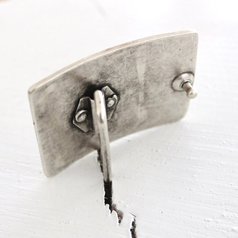 May include: A silver belt buckle with a textured surface and a hinged closure. The buckle is rectangular in shape and designed to be attached to a belt.