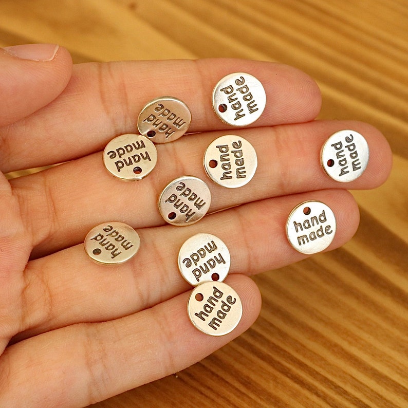 K&ouml;nnte beinhalten: Nahaufnahme von zehn goldfarbenen Metall-Etiketten mit der Aufschrift "hand made" auf jeder. Die Etiketten sind rund und haben ein Loch in der Mitte.